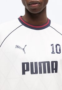 Κοντινό πλάνο ατόμου που φοράει λευκό πουκάμισο Puma με λεπτομέρειες κόκκινου και μπλε στο γιακά, λογότυπο Puma στο αριστερό στήθος και αριθμό 10 στο δεξί στήθος.