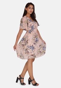 Blumenkleid in hellem Rosa mit kurzen, fließenden Ärmeln, elastischem Bund und schwingendem Rock. Mit blauen und lila Rosen sowie grünen Akzenten. Schwarze Absatzsandalen.