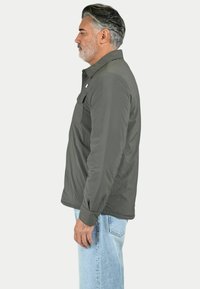 Giacca isolante grigia con collo a camicia, tasca frontale e maniche lunghe, indossata su jeans azzurri chiari. Texture liscia e design casual.