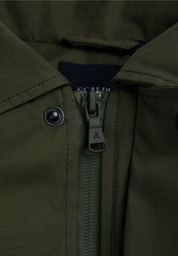 Chaqueta verde oliva con cierre frontal de cremallera, botones a presión y tejido texturizado. Incluye una etiqueta y un tirador de cremallera en contraste.