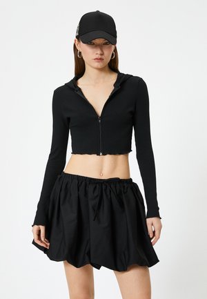 Schwarze, verkürzte Kapuzenjacke mit Reißverschluss und gerippter Textur, kombiniert mit einem schwarzen, gerafften Rock mit elastischem Bund und Kordelzug.