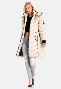 Puffige beige Jacke mit hohem Kragen, schwarzen Akzenten und vorderem Reißverschluss. Verfügt über eine gep quiltete Textur und ein Patch auf dem linken Ärmel.
