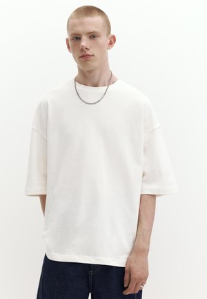 Mlad moški s kratkimi lasmi, ki nosi preveliko belo majico, temne kavbojke, verižico in prstan na desni roki.