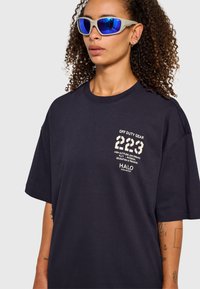 Marinblå oversize T-shirt med tryckt text i krämfärg, med rund halsringning och korta ärmar. Modellen bär blåtonade solglasögon.