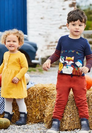 Deux tout-petits se tiennent près de bottes de paille en plein air ; une fille en robe jaune et un garçon en chemise colorée et pantalon rouge portent tous deux des bottes noires.