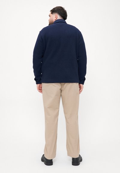 Ναυτικό φλις μπουφάν με γιακά, μακριά μανίκια, χαλαρή εφαρμογή, συνδυασμένο με μπεζ παντελόνι και μαύρα παπούτσια, από πίσω.