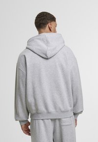 Uomo di spalle che indossa una felpa grigia chiara con cappuccio e pantaloni abbinati, su uno sfondo bianco semplice.