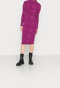 Robe ajustée à manches longues de couleur rose vif agrémentée d'un motif en vagues noires, accompagnée de bottines en cuir noir à semelle épaisse. Texture lisse, longueur mi-mollet.