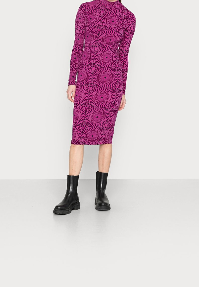 Robe ajustée à manches longues de couleur rose vif agrémentée d'un motif en vagues noires, accompagnée de bottines en cuir noir à semelle épaisse. Texture lisse, longueur mi-mollet.