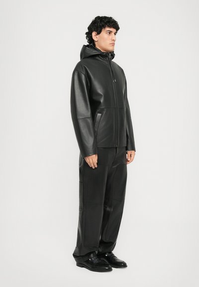 Bărbat în picioare, îmbrăcat într-o jachetă cu glugă din piele neagră, pantaloni din piele asortați și pantofi eleganți negri, pe un fundal simplu, deschis.