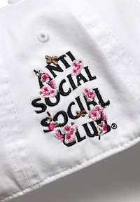 Biała czapka baseballowa z czarnym haftowanym napisem "ANTI SOCIAL SOCIAL CLUB" otoczonym różowymi i brązowymi motywami kwiatowymi.