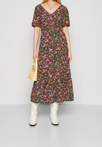 Sukienka maxi w kwiaty w kolorze czarnym z różowymi, pomarańczowymi i zielonymi wzorami, dekolt w kształcie litery V, krótkie rękawy, zestawiona z białymi botkami i żółtą torebką.