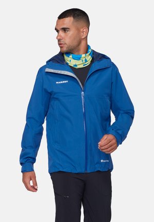 Homme portant une veste imperméable bleue avec capuche, un pantalon noir et un tour de cou bleu-jaune à motifs, regardant vers sa gauche.