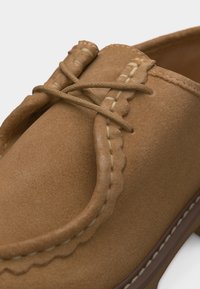 Mocassin en daim marron clair avec coutures mocassin et un nœud décoratif à l'avant, contrasté par une semelle marron plus foncée.