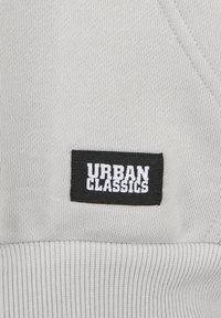 Etichetta in tessuto rettangolare nera con testo bianco "URBAN CLASSICS" su tessuto a coste di un grigio chiaro.