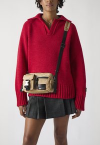 Red knit sweater with a v-neck and ribbed cuffs, paired with a beige crossbody bag featuring a zip pocket and silver hardware on a plaid skirt.
