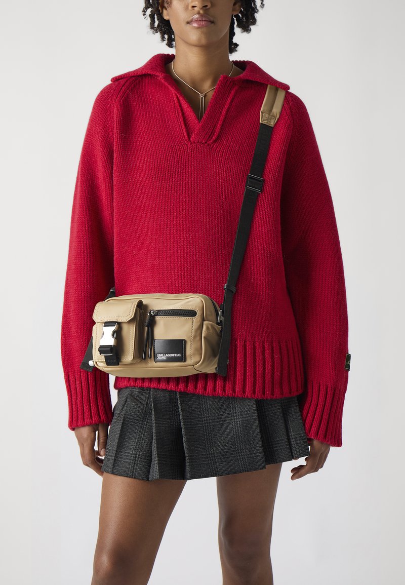 Red knit sweater with a v-neck and ribbed cuffs, paired with a beige crossbody bag featuring a zip pocket and silver hardware on a plaid skirt.