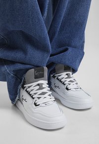 Baskets blanches avec des accents gris, au design montante et en matériau texturé. Présente un logo noir sur le côté et des lacets blancs. Portées avec un jean en denim ample.