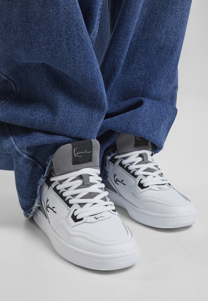 Baskets blanches avec des accents gris, au design montante et en matériau texturé. Présente un logo noir sur le côté et des lacets blancs. Portées avec un jean en denim ample.