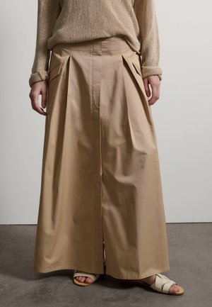 Persona con un suéter beige de punto, pantalones caqui de tiro alto y pierna ancha con pliegues, y sandalias crema, de pie sobre un suelo gris.