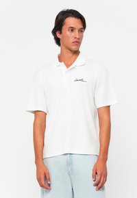 Polo blanc à manches courtes en mélange de coton avec une coupe décontractée, présentant un logo en cursive noir sur la poitrine et un col classique.
