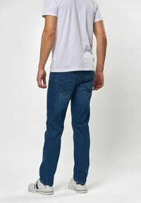 Kék farmernadrág egyenes szabással, hátsó zsebekkel, minimális varrással és enyhe kifakulással; egyszerű fehér pólóval és tornacipővel viselve.