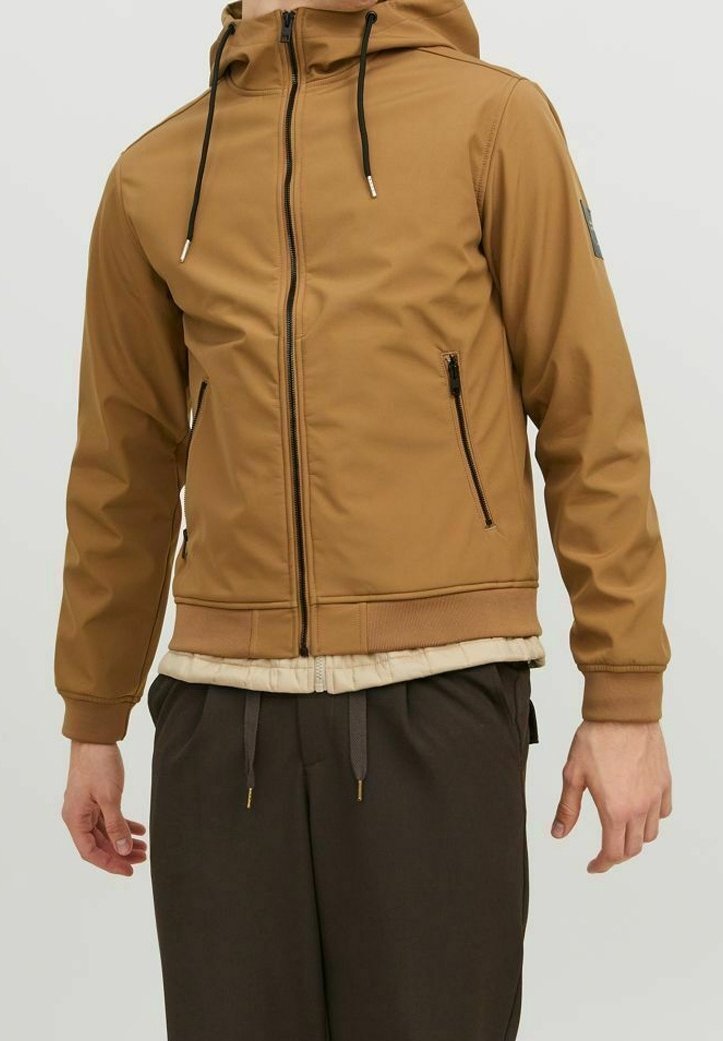Homme portant une veste à capuche beige zippée avec des cordons noirs et des poches zippées, associée à un pantalon foncé.