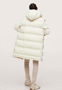 Gilet sans manches rembourré en ivoire avec capuche. Rayures horizontales texturées sur toute la surface. Porté sur un pantalon clair à côtes et des chaussures.