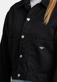 Chaqueta de mezclilla negra con botones a presión, con un parche del logo en forma de corazón en el bolsillo. Diseño estructurado con dos bolsillos frontales.