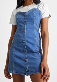 Robe en denim avec des bretelles spaghetti, délavée claire, fermeture à boutons sur le devant, design ajusté et coutures latérales, portée par-dessus un t-shirt blanc.