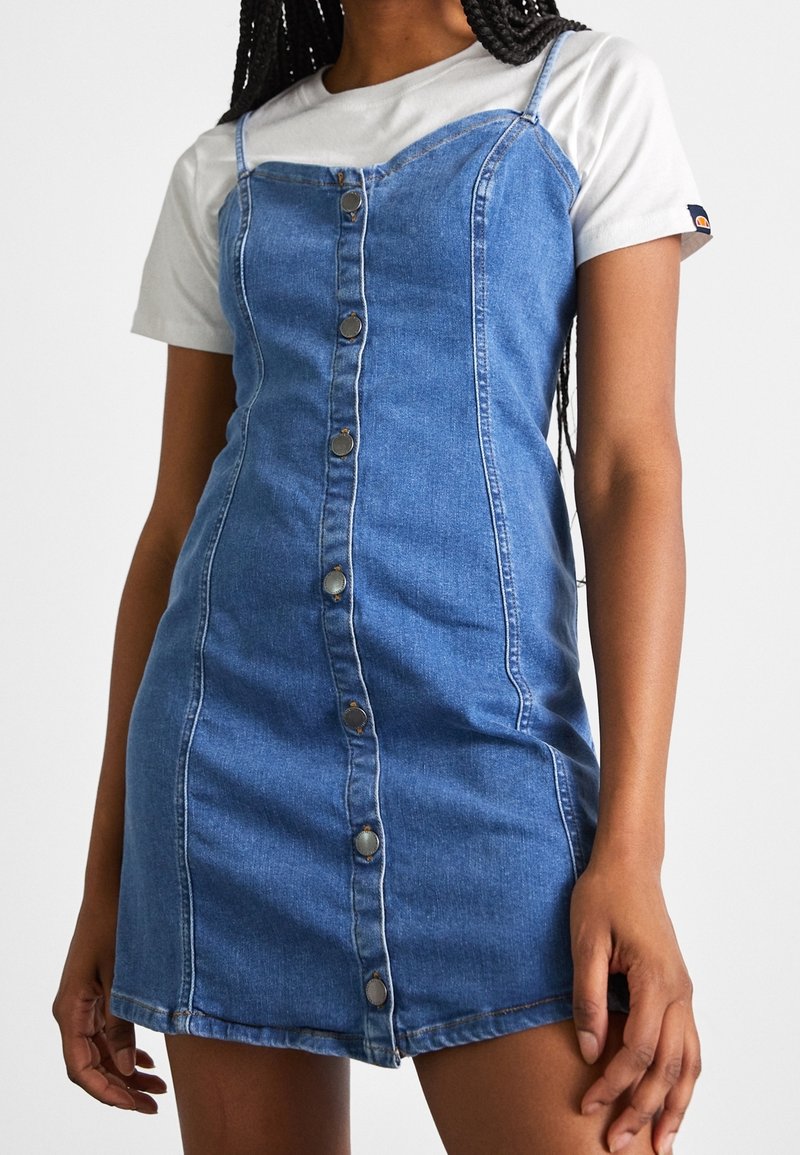 Robe en denim avec des bretelles spaghetti, délavée claire, fermeture à boutons sur le devant, design ajusté et coutures latérales, portée par-dessus un t-shirt blanc.