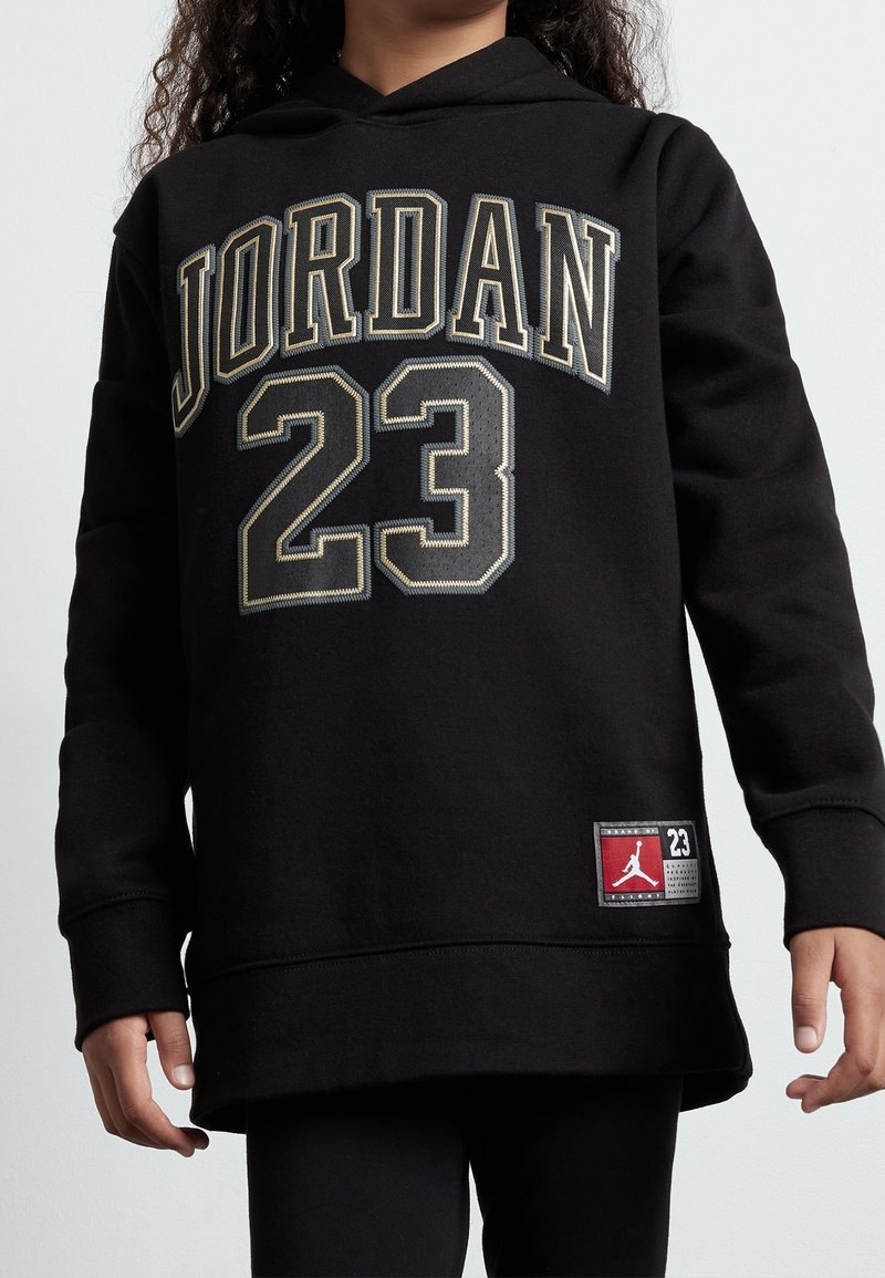 Černá mikina s kapucí má ve zlaté nitě nápis "JORDAN" a číslo "23". Na lemu je logo nášivka s červeným a černým designem.