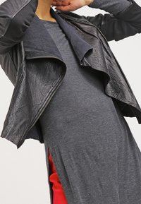 Giacca in pelle nera con colletto asimmetrico, finitura testurizzata e chiusura con zip, indossata sopra un vestito grigio a maniche lunghe con uno spacco laterale rosso.