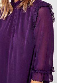 Blusa viola con scollo e maniche a volants. Tessuto trasparente con una texture leggera e polsini aderenti, che conferisce un aspetto morbido e fluente.