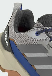 Šedá sportovní obuv má texturovaný síťovaný svršek, modré a hnědé akcenty a polstrovanou krémovou podrážku s poutkem na patě.