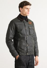 Homme portant une veste vert foncé avec des poches avant boutonnées et un écusson sur la poitrine, associé à un jean sombre, se tenant devant un arrière-plan uni.