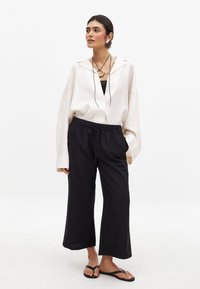 Lichtbeige oversized blouse met ingesneden kraag, gecombineerd met losse zwarte broeken en zwarte slippers. Eenvoudige ketting benadrukt de look.