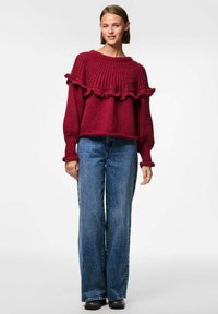 Suéter rojo de punto con cuello acanalado y dobladillo con volantes, combinado con unos pantalones vaqueros de mezclilla azul de cintura alta y zapatos negros.