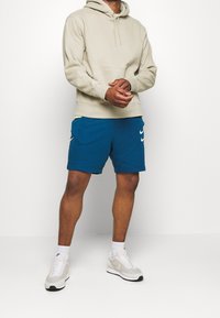 Light beige hoodie with a front pocket, paired with blue shorts featuring white Nike logos. White and grey sneakers, white socks.