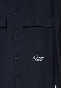 Chemise boutonnée bleu marine en tissu à texture lisse, avec deux poches poitrine et texte brodé blanc en bas à droite.