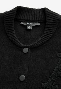 Zwarte gebreide cardigan met een geribbeld kraag, voorzien van twee zwarte knopen en een gestructureerde letter "A" detail op de voorkant. Maatlabel zichtbaar.