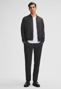 Giacca bomber nera in tessuto testurizzato con polsini a costine, t-shirt bianca sotto e pantaloni aderenti scuri abbinati. Scarpe nere con lacci completano il look.