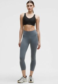 Sujeción deportiva negra y leggings ajustados grises, con una cinturilla alta, textura suave y corte a la altura de la rodilla, combinados con zapatillas deportivas ligeras.
