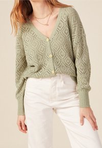 Jasnozielony dziany kardigan z wzorem w diamenty, z dekoltem w serek, ściągaczami na mankietach i żółtymi guzikami. Do tego jasne spodnie.