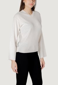 Maglione bianco a V con texture a rombi e polsini a coste, abbinato a pantaloni neri. Il maglione ha maniche lunghe e una vestibilità comoda.