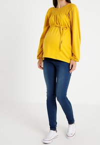 Gul graviditetsblus med smockad halsringning och långa ärmar, knuten i midjan, kombinerad med mörkblå tighta jeans och vita sneakers.