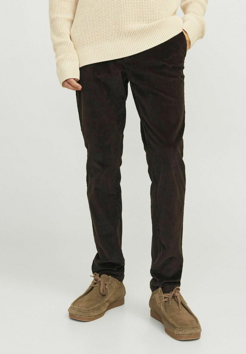 Pantalon en velours côtelé marron foncé avec une coupe slim, un tissu texturé et de légères rayures verticales, associé à des chaussures en daim marron clair.