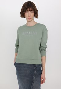 Zelená mikina s žebrovanými manžetami a lemem, na přední straně s nápisem "ARMANI EXCHANGE" v lesklých stříbrných písmenech.