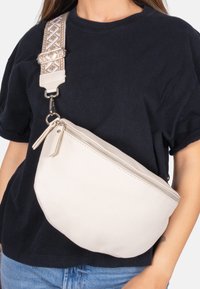 Cremfarbene Leder-Crossbody-Tasche mit strukturierter Oberfläche, geschwungener Form und silbernem Reißverschluss. Verstellbarer Riemen mit geometrischem Muster.