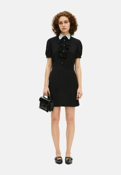 Rochie scurtă neagră cu mâneci bufante, față frufru și închidere cu nasturi. Guler din dantelă albă. Modelul ține o poșetă mică neagră și poartă pantofi cu talpă joasă negri.